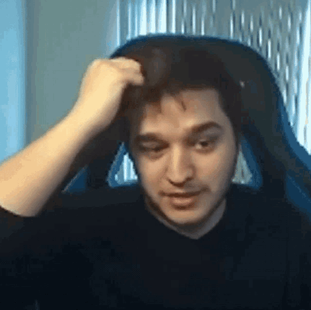 a man in a black shirt is sitting in a blue chair and scratching his head .