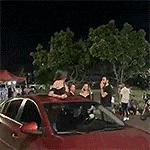 a group of people are standing around a red car in a parking lot .