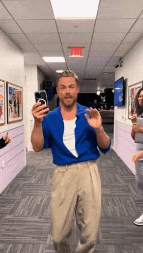 a man in a blue shirt is taking a picture of himself in a hallway with a red exit sign above him