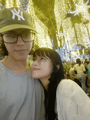 a man wearing a ny hat stands next to a woman with glasses
