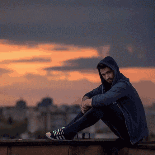 a man wearing a hoodie sits on the edge of a building at sunset