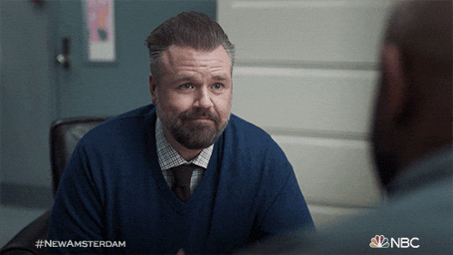 a man in a blue sweater and tie is talking to another man with the nbc logo behind him