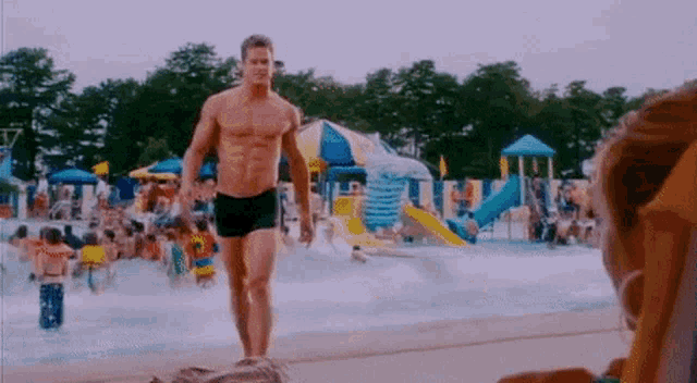 a shirtless man is walking through a water park while a woman sits in a chair .