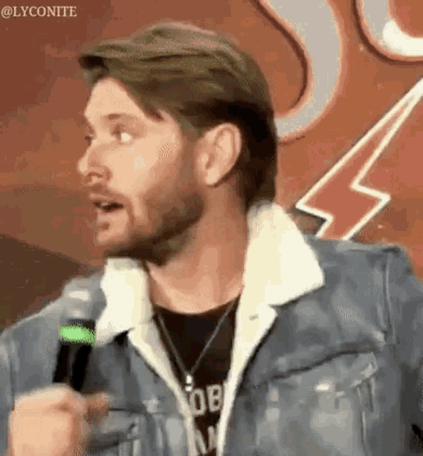 a man with a beard is talking into a microphone while wearing a denim jacket .