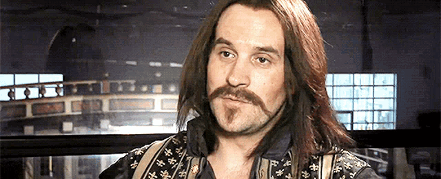 a man with long hair and a mustache is wearing suspenders and a shirt with chinese characters on it