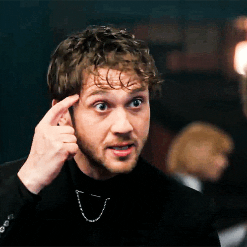 a man wearing a black turtleneck and a chain around his neck holds his finger to his forehead