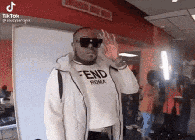 a man wearing sunglasses and a fendi roma shirt is waving