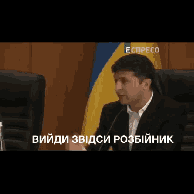 a man in a suit speaking into a microphone with expresso in the corner