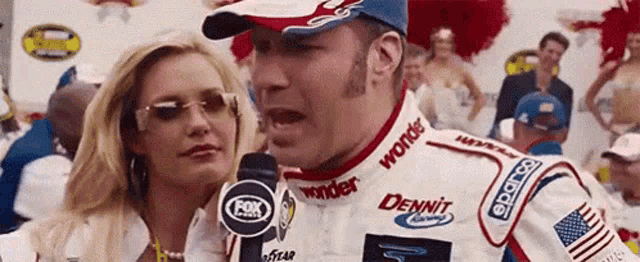 a man in a race car jacket is talking into a microphone while a woman looks on .