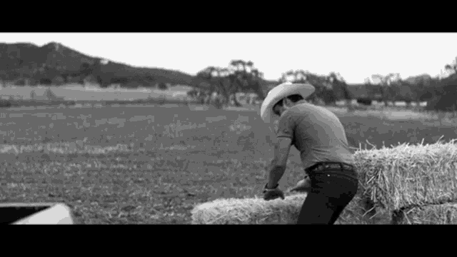 a black and white photo of a man in a cowboy hat pushing a bale of hay in a field .