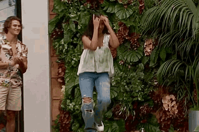 a woman is dancing in front of a wall of plants while a man applauds .