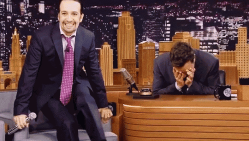 a man in a suit and tie is sitting in a chair holding a microphone and laughing .