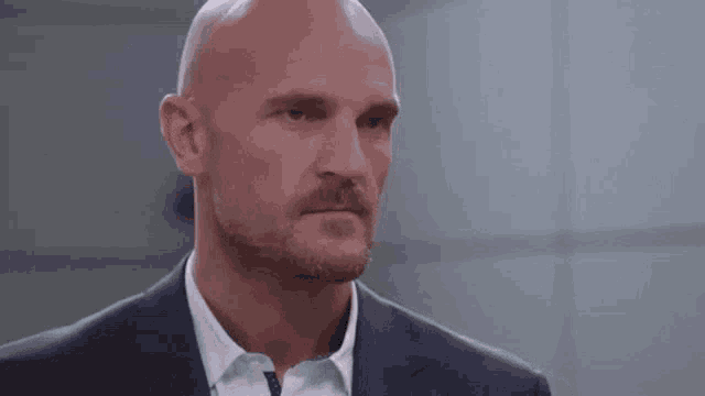 a bald man with a beard is wearing a suit and white shirt and looking at the camera .