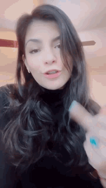 a woman with long dark hair is wearing a black turtleneck and a blue nail polish