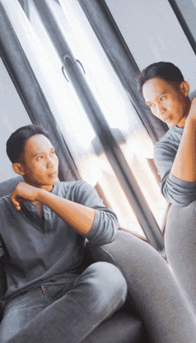 a man sitting in front of a window looking at his reflection in the mirror