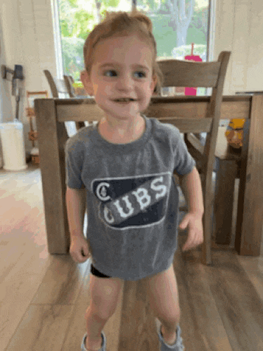 a little girl wearing a cubs t-shirt stands in front of a wooden table