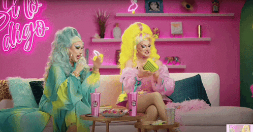two drag queens are sitting on a couch in front of a neon sign that says ' so to diego '