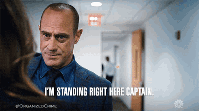 a man in a suit and tie says " i 'm standing right here captain " in a hallway