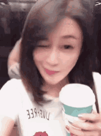 a woman winks while holding a cup of coffee and wearing a white shirt that says dae-eee