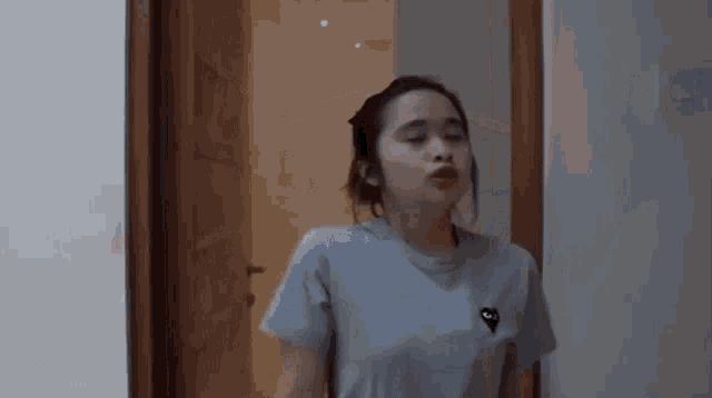 a woman in a blue t-shirt is standing in front of a door and making a funny face .