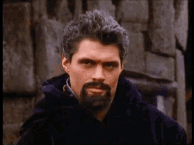 a man with a beard and gray hair is wearing a black cape and standing in front of a stone wall .