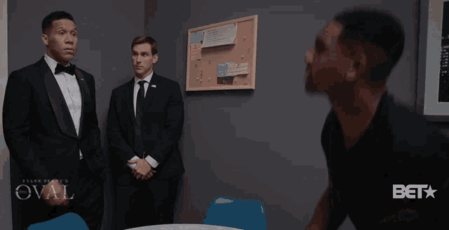 a man in a tuxedo stands next to a man in a black shirt and says sit