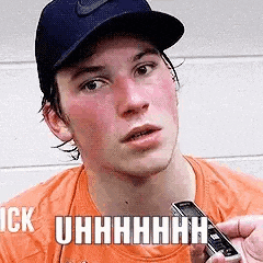 a young man wearing a hat and an orange shirt is holding a cell phone and making a funny face .