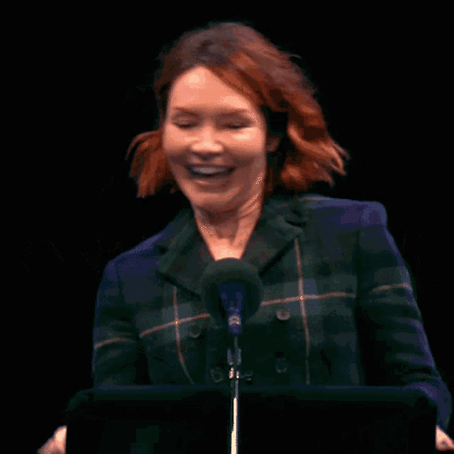 a woman in a plaid jacket stands in front of a microphone with her hands in the air