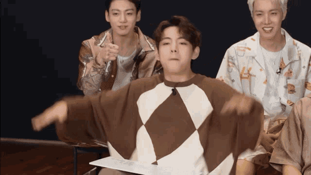 a group of young men are sitting around a table and one of them is wearing a brown and white sweater with a checkered pattern
