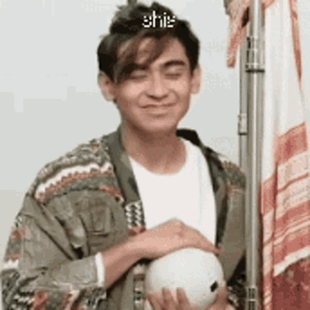 a young man is holding a soccer ball and smiling while wearing a green jacket and a white shirt .