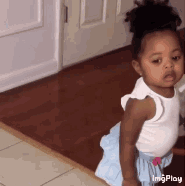 a little girl in a white tank top and blue skirt is standing on a tiled floor in front of a door .
