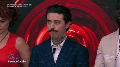 a man with a mustache and a tattoo on his neck stands in front of a red background that says #grandefratello