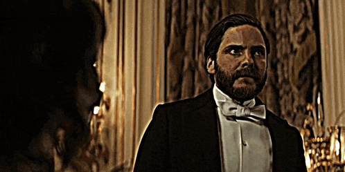 a man with a beard wearing a tuxedo and bow tie talks to another man