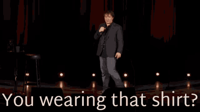 a man in a suit is speaking into a microphone with the words " you wearing that shirt " below him