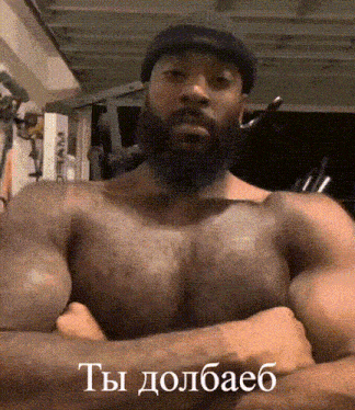 a shirtless man with a beard is standing with his arms crossed and says " ты долбаеб "