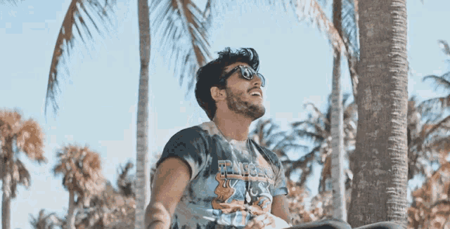 a man wearing sunglasses and a t-shirt that says peace on it sits under a palm tree
