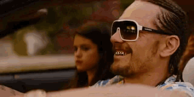 a man wearing sunglasses is driving a car with a girl in the back .