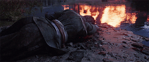 a man is laying on the ground in front of a river with a fire in the background