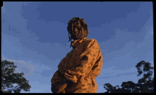 a man with dreadlocks stands in front of a blue sky with trees in the foreground
