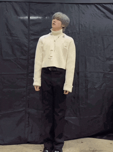 a man wearing a white sweater and black pants stands in front of a black tarp