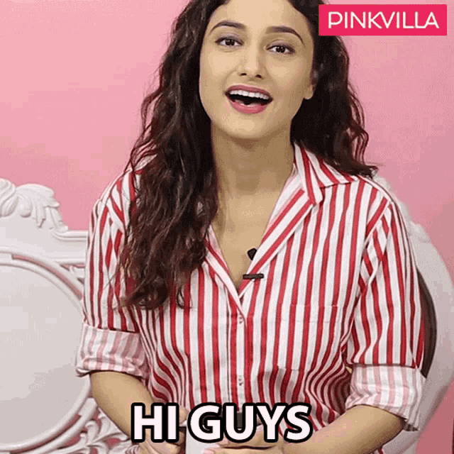 a woman wearing a red and white striped shirt says hi guys in front of a pink wall