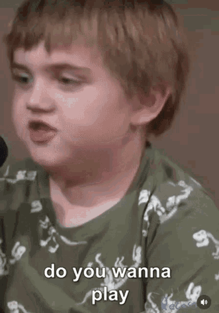 a young boy is talking into a microphone while wearing a green shirt that says `` do you wanna play '' .