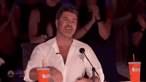 a man in a white shirt is sitting at a table in front of a microphone and drinking a dunkin donuts drink .