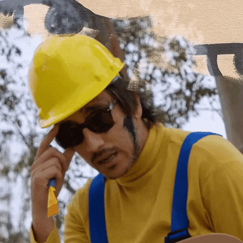 a man wearing a yellow hard hat and sunglasses holds his hand to his forehead