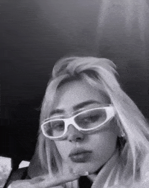 a black and white photo of a woman wearing glasses and making a peace sign .