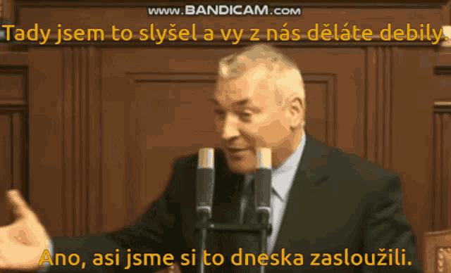 a man in a suit and tie stands in front of two microphones with the words www.bandicam.com in the corner