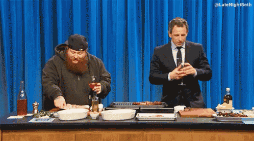 a man in a suit stands next to a man with a beard in front of a blue curtain that says latenightseth
