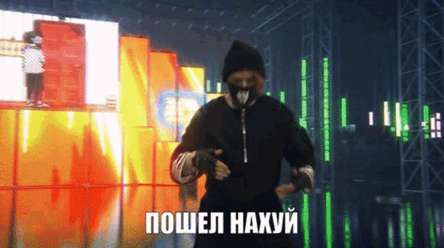 a man in a mask is dancing in front of a large screen that says " пошел нахуи "