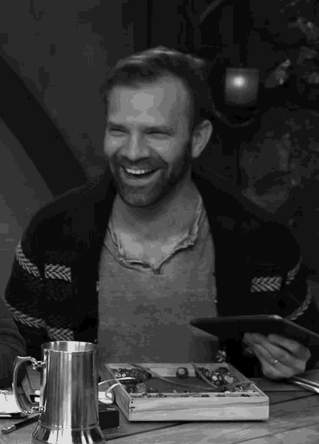 a man with a beard is smiling while sitting at a table with a silver mug
