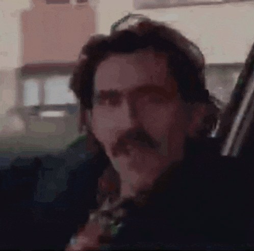 a man with a mustache is holding a gun in a car and looking at the camera .